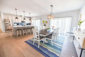 A modern kitchen with stainless steel appliances, a dining area with a wooden table and white chairs, and a large window with sheer curtains.