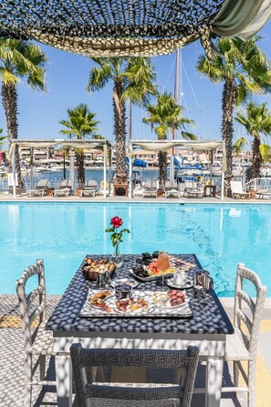 A poolside dining area with a table set for a meal, surrounded by lounge chairs and palm trees. - Ianos Bay (Lefkada)