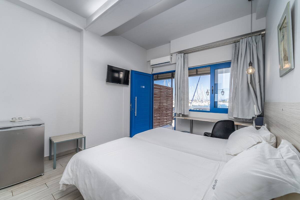 Ett hotellrum med en säng, ett skrivbord, en stol, en tv och ett fönster med utsikt över båtar.