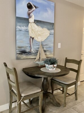 A dining area with a round table, two chairs, a potted plant, and a painting of a woman by the sea.