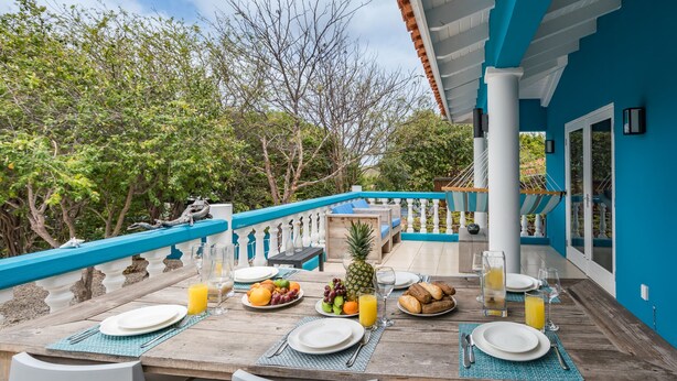 A dining table set with plates, cutlery, and a variety of fruits and beverages, surrounded by white chairs, with a blue railing and trees in the background. - Villa Despacito (Sint Willibrordus)