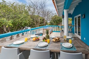 Een eettafel gedekt met borden, bestek en diverse soorten fruit en drankjes, omringd door witte stoelen, met een blauwe balustrade en bomen op de achtergrond.