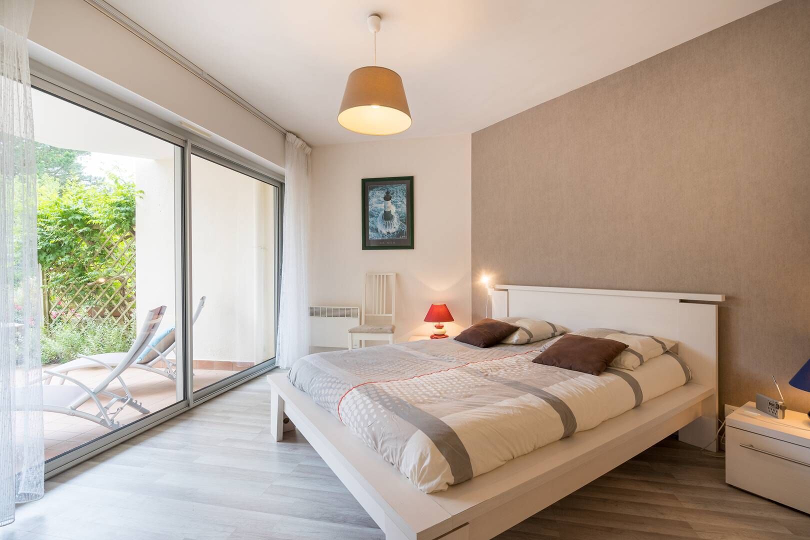A modern bedroom with a large bed, a bedside table, a chair, and a view of a garden through a sliding glass door.