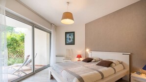 A modern bedroom with a large bed, a bedside table, a chair, and a view of a garden through a sliding glass door. - Carnac - 3-room apartment - 69m² - Wifi (CARNAC)