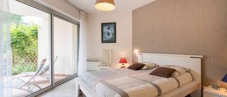 A modern bedroom with a large bed, a bedside table, a chair, and a view of a garden through a sliding glass door.