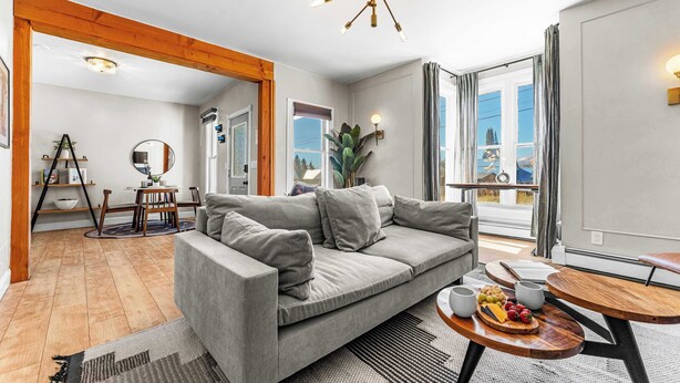 A modern living room with a grey sofa, wooden beams, a dining area with a table and chairs, and a large window with curtains. - Pine Street Retreat | 1BR Hot Tub Home, Dog Door, Walk to Main St | Leadville (Leadville)