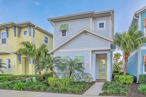 A two-story building with a yellow facade and a blue roof, surrounded by greenery and palm trees.