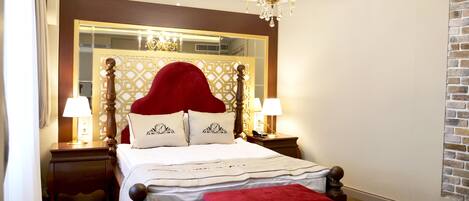 A bedroom with a large bed, two bedside tables, a red upholstered bench, a chandelier, a mirror, and a brick-patterned wall.