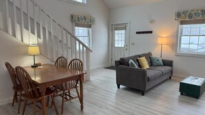 A spacious living room with a wooden dining table and chairs, a gray sofa with cushions, and a staircase with white railings. - Western Mountain Townhouse (Southwest Harbor)