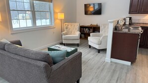 A living room with a grey sofa, two armchairs, a coffee table, a TV mounted on the wall, and a kitchen area with a stove and cabinets. - Western Mountain Townhouse (Southwest Harbor)