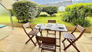 A patio with a table set for four, surrounded by wooden chairs, overlooking a garden with various plants and a white building in the background. - Etel - 2-room apartment - 40m² - Wifi - 3* (ETEL)