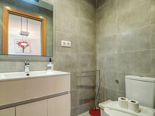 Un baño moderno con un lavamanos, espejo y retrete blancos. Hay una zona de ducha con puerta de cristal y una alfombra roja.