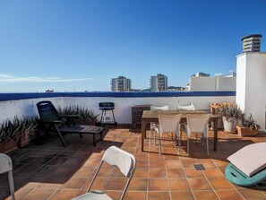 Une terrasse sur le toit offrant une vue sur la mer et les bâtiments, avec une table, des chaises et des plantes en pot.