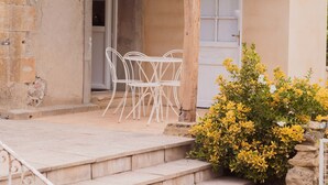 Un bâtiment en pierre avec un toit en tuiles, une pergola en bois, une porte blanche et un petit jardin avec des fleurs jaunes.
