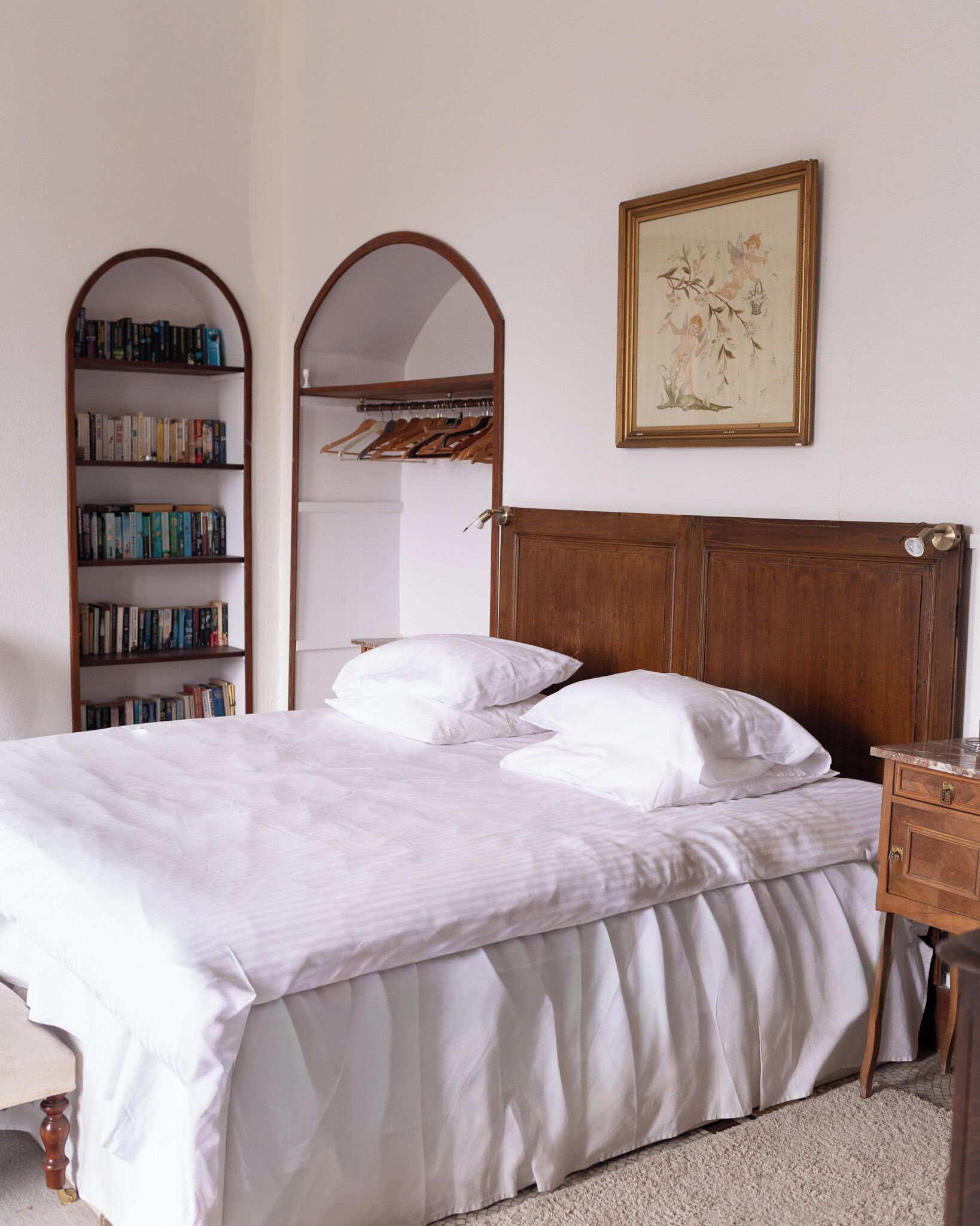 A neatly made bed with white linens, a wooden headboard, and a bedside table with a lamp. There is a bookshelf filled with books and a framed picture on the wall.