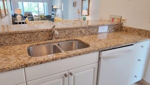 Una cocina con encimeras de granito, un lavamanos de acero inoxidable, armarios blancos y un comedor visible al fondo.
