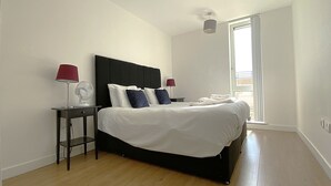 A bedroom with a large bed, a bedside table with a lamp, a fan, and a window with a view.