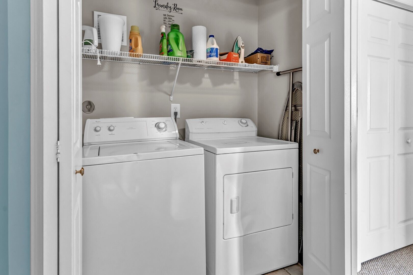 A laundry room with two white front-loading washing machines, a shelf with various cleaning products, and a door to the right.