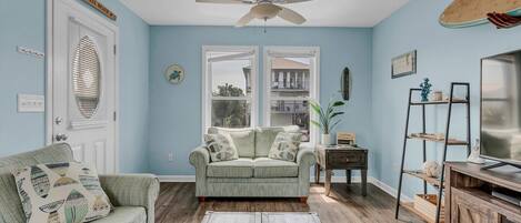 Un salon avec un mur bleu clair, un ventilateur de plafond, un canapé agrémenté de coussins à motifs, une table basse avec des télécommandes et un meuble TV en bois.