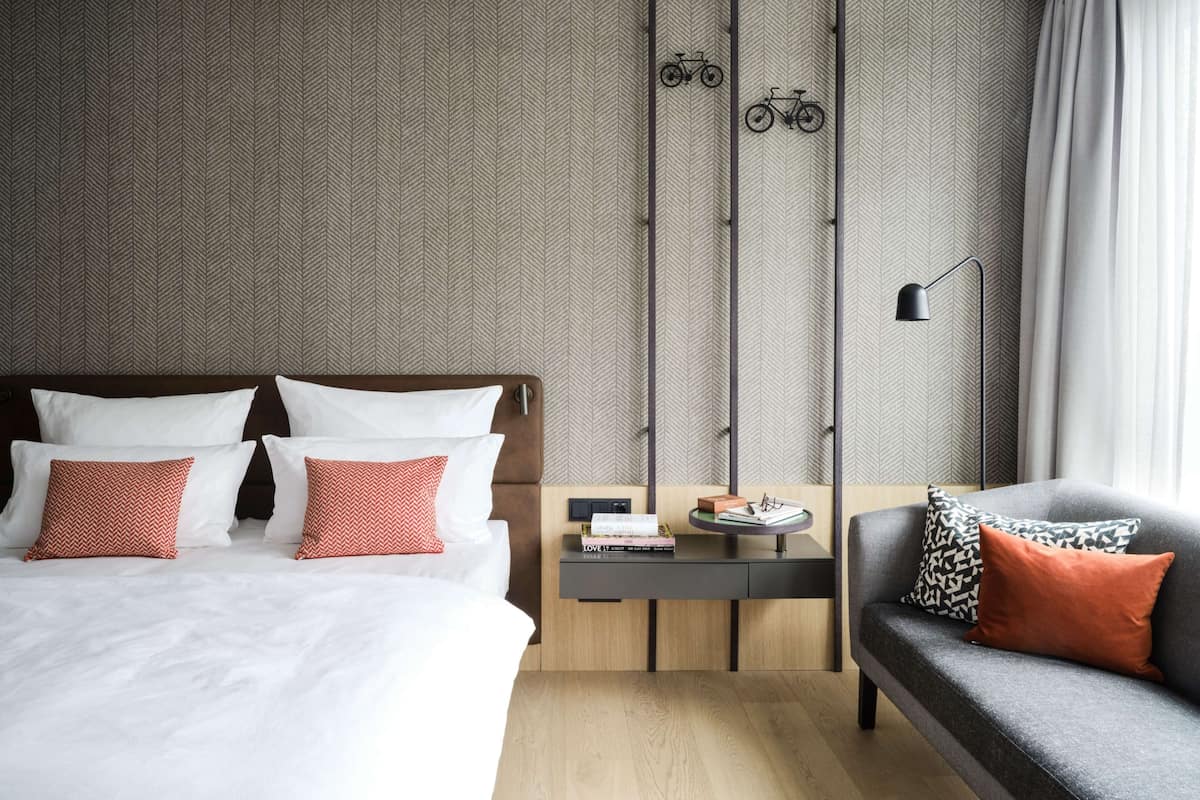 A hotel room with a bed, a nightstand, a sofa, and a wall with a herringbone pattern.