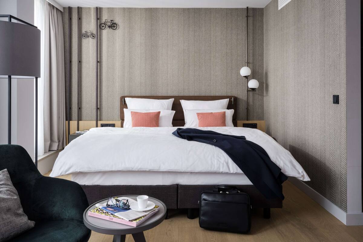 A modern hotel room with a large bed, a bedside table, a chair, a small table with a magazine and glasses, and a suitcase.