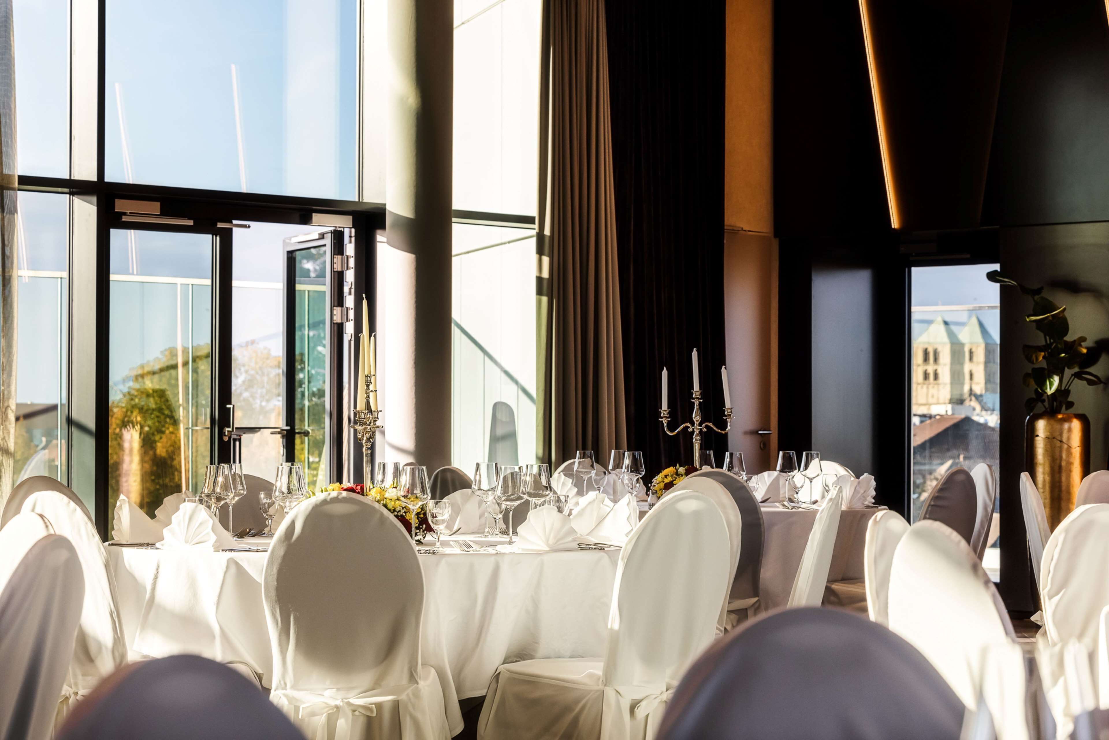 A banquet hall with round tables set for a formal event, large windows with a view of buildings, and a modern interior design.