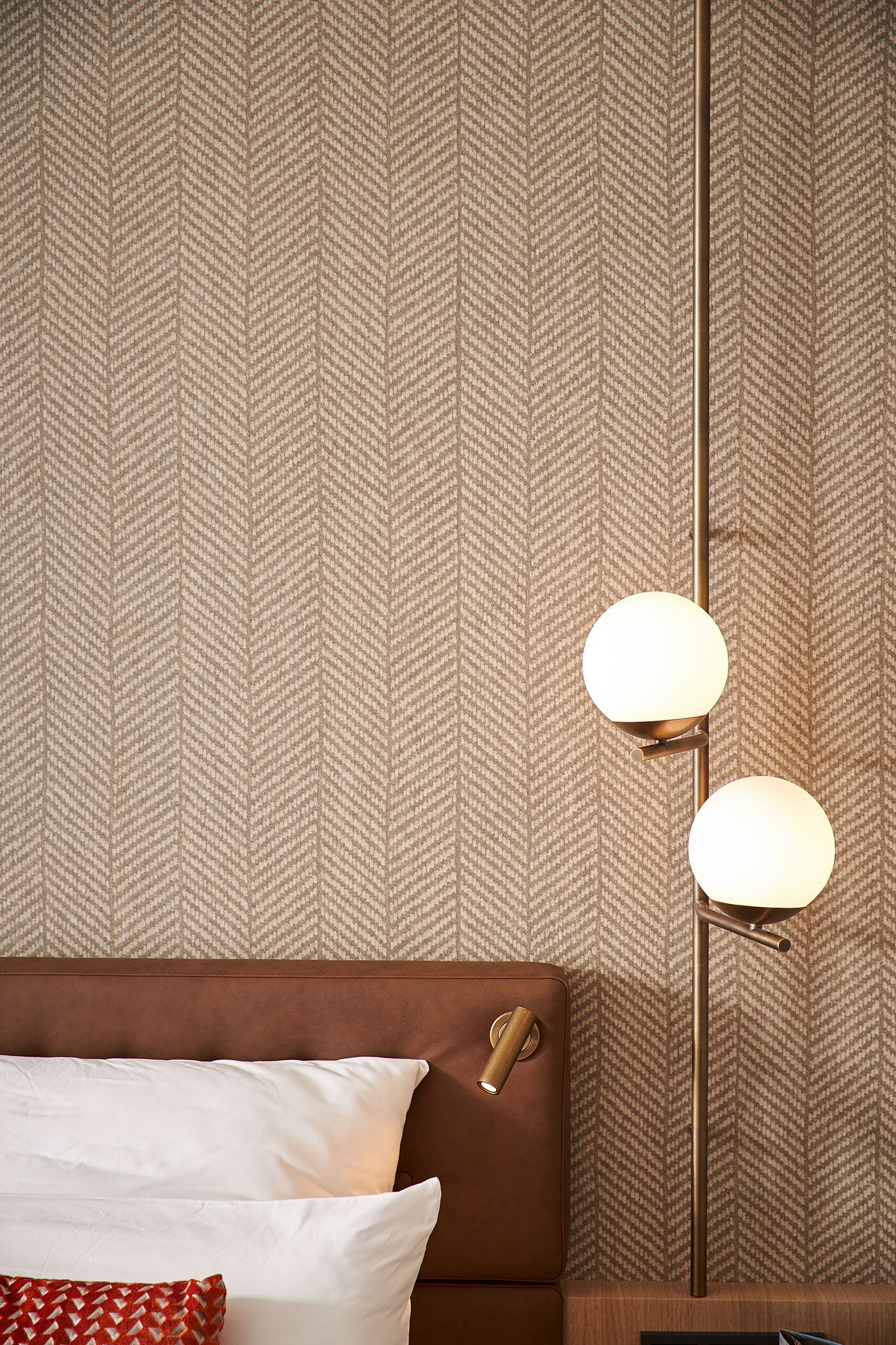 Une chambre d’hôtel avec un papier peint à motifs, une tête de lit marron, une table de chevet avec une lampe et un oreiller rouge et blanc.