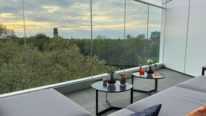 Un balcón moderno con vista a un paisaje urbano, que incluye una torre y dos pequeñas mesas con adornos.