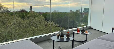 Un balcón moderno con vista a un paisaje urbano, que incluye una torre y dos pequeñas mesas con adornos.