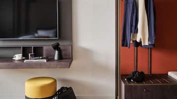 Una habitación de hotel moderna con una televisión de pantalla plana, una mesa de centro con una taza, libros, un Cubo de Rubik, una lámpara, una maleta, un banco con zapatos y un perchero con varias prendas de vestir.