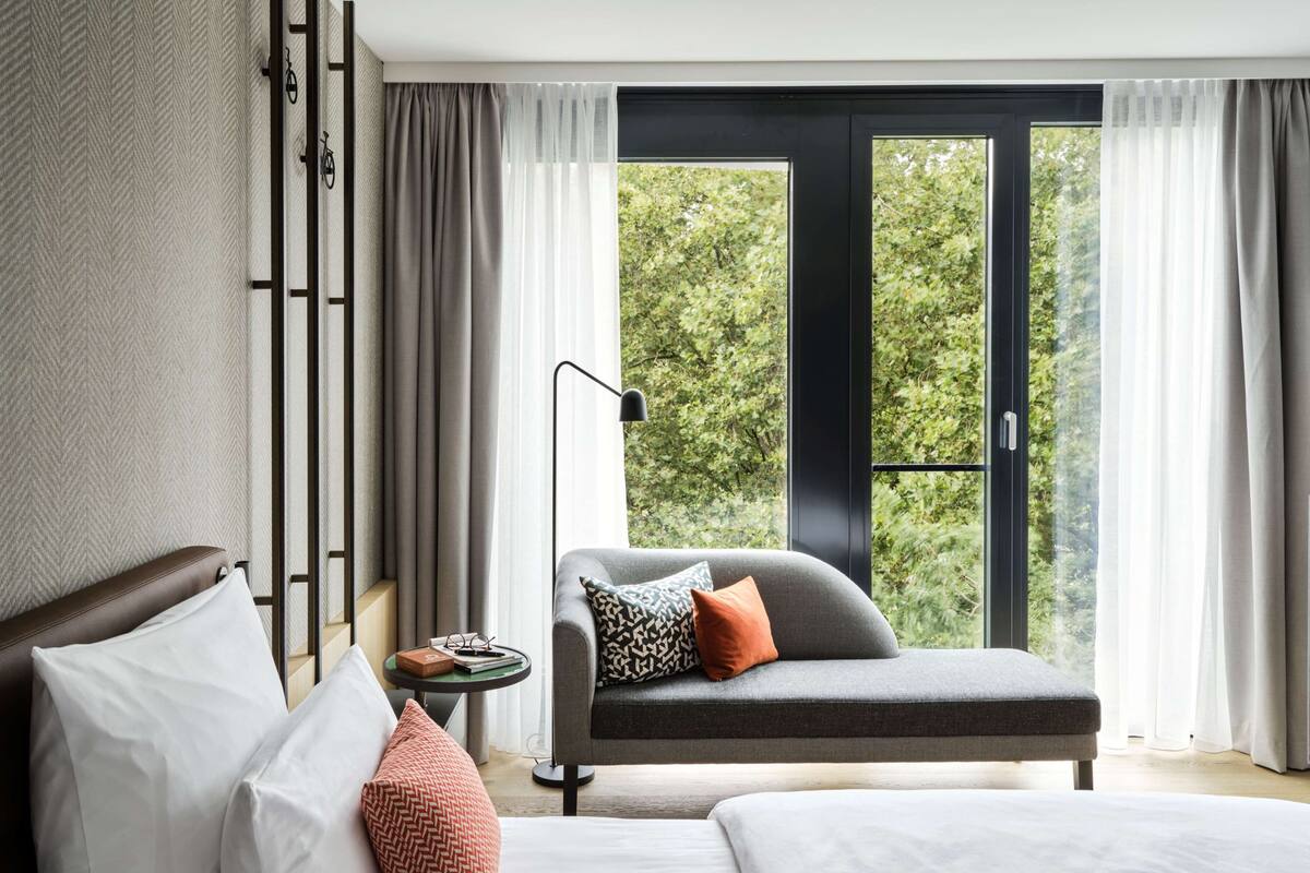 A modern hotel room with a bed, a sofa, a small table with a lamp, and a view of greenery through large windows.