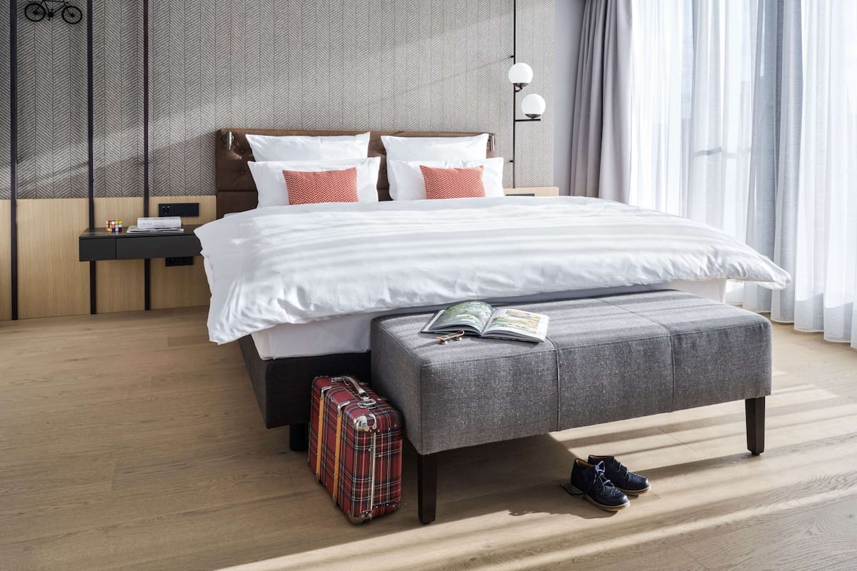 A modern hotel room with a large bed, a bedside table with a lamp, a suitcase, and a pair of shoes on the floor.