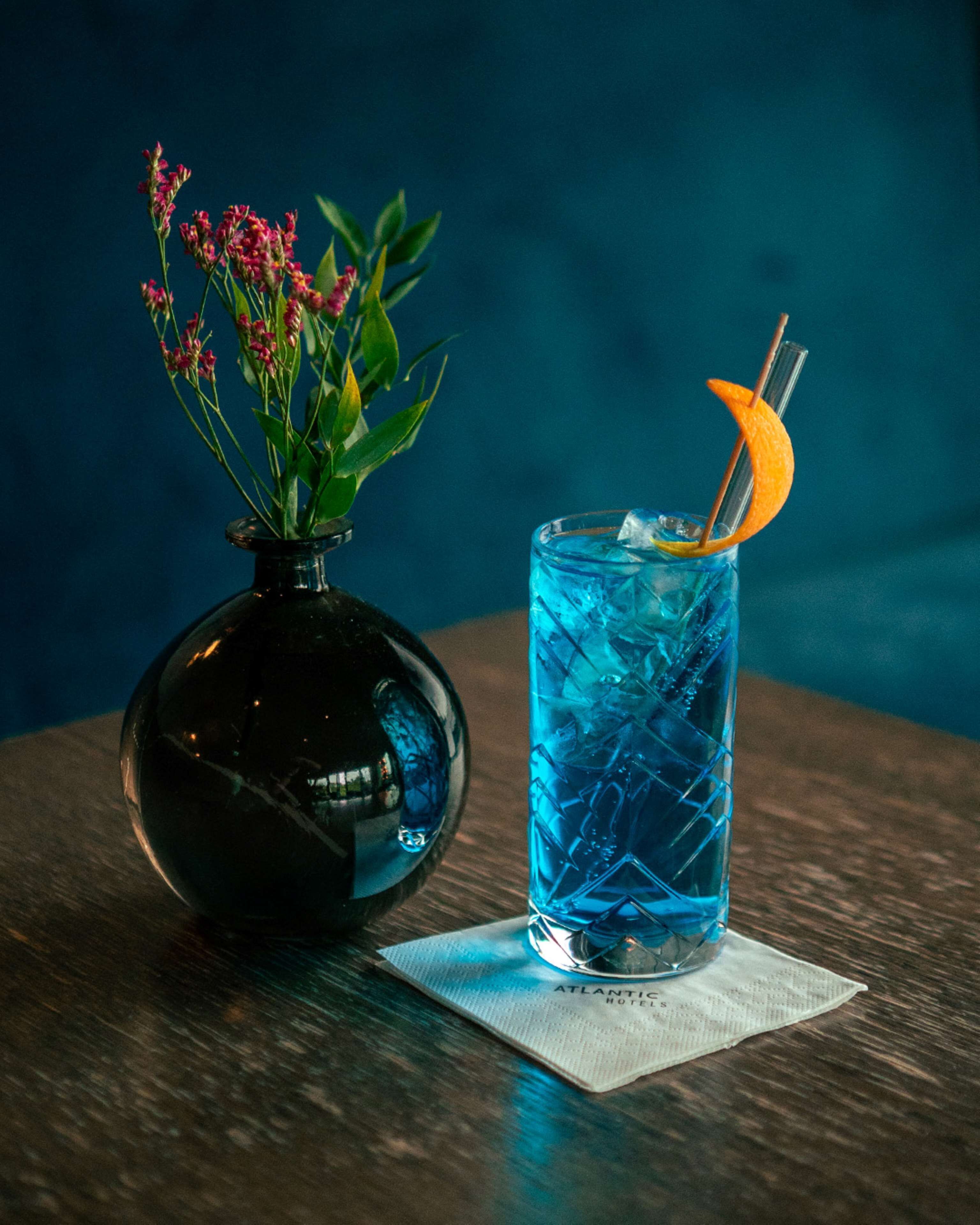 A dark blue glass with a lemon slice and a straw, placed on a wooden table next to a black vase with pink flowers.