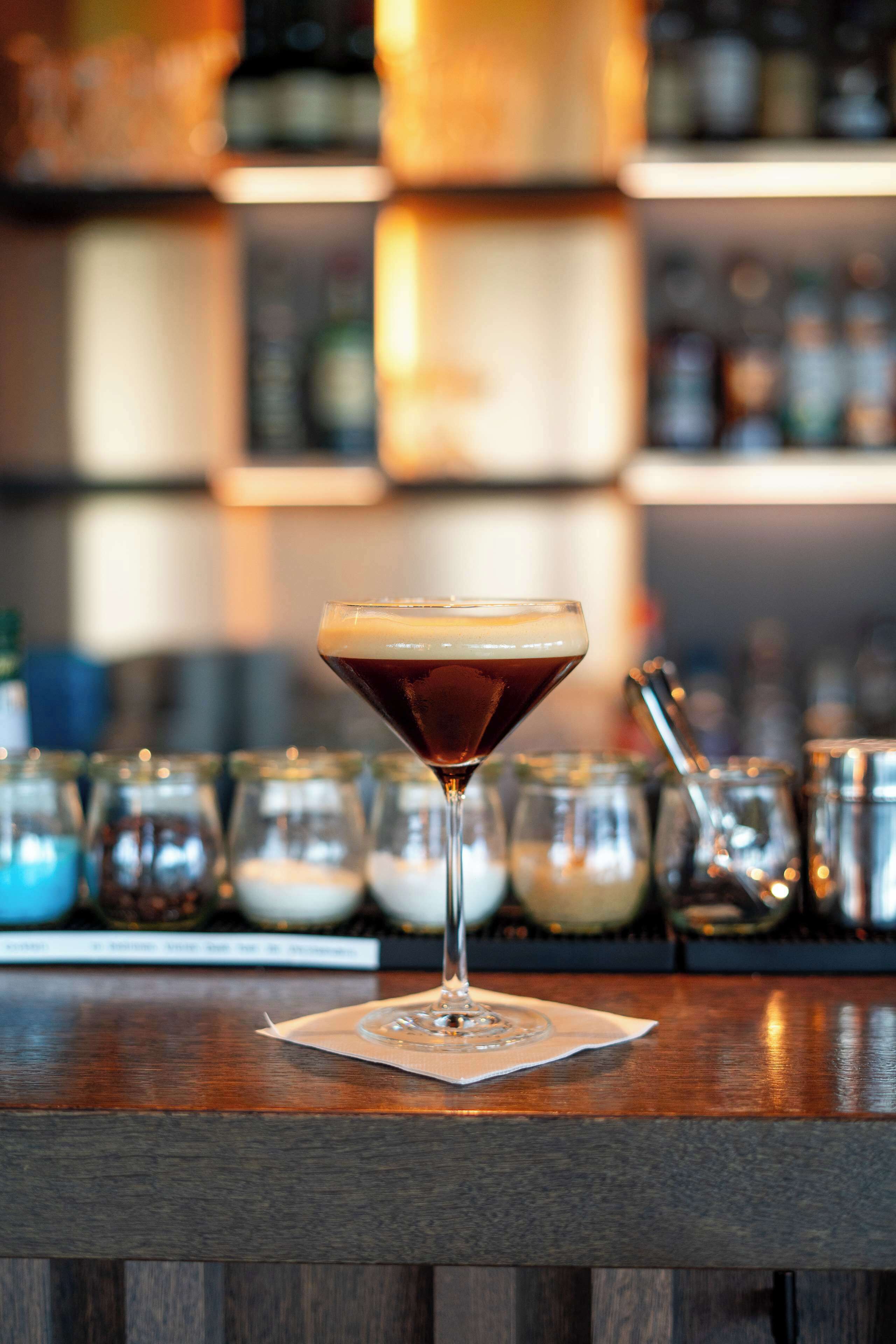 Un verre à martini, surmonté d’une mousse, est posé sur le comptoir d’un bar, entouré de divers ustensiles et bouteilles de bar.
