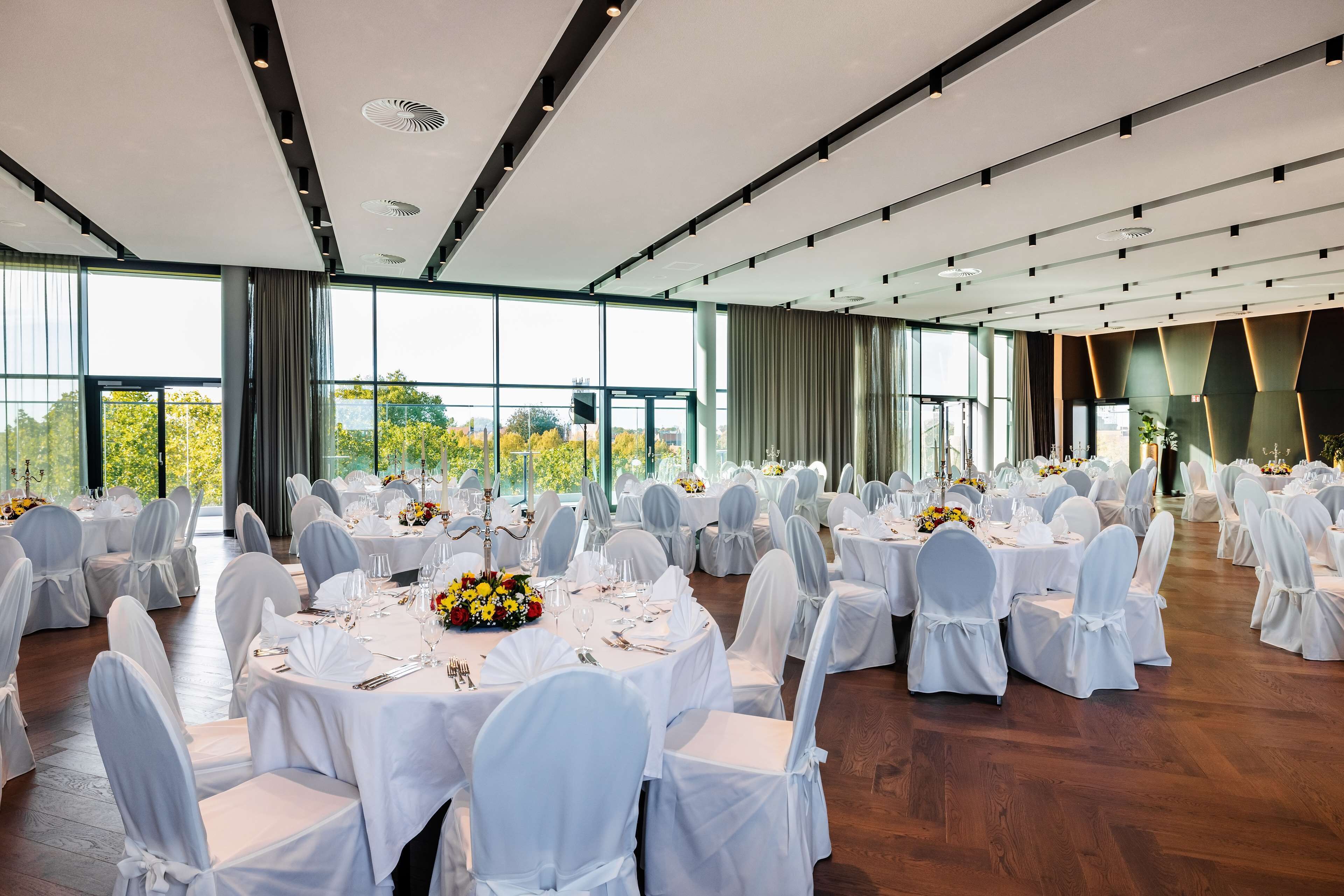 Une salle de banquet avec des tables rondes dressées pour le repas, des chaises blanches et de grandes fenêtres donnant sur un paysage verdoyant.