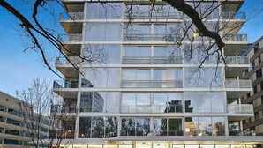 Um edifício moderno de vários andares, com grandes janelas e varandas, rodeado por árvores e outros edifícios.