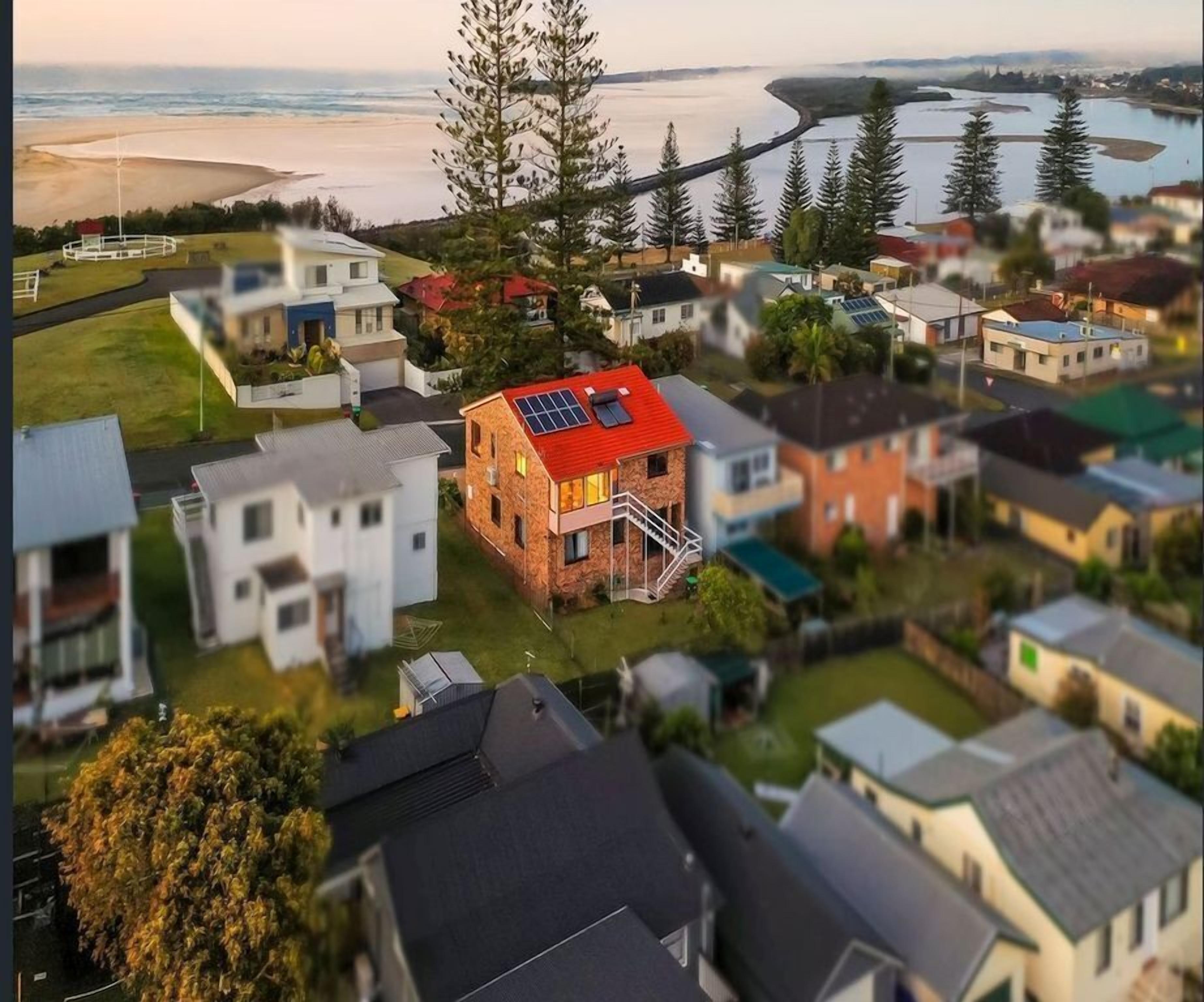 Zona residencial con casas, una vivienda destacada con techo rojo y paneles solares, y una playa al fondo.