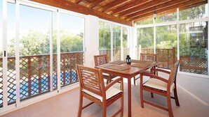 A covered patio with a wooden table and chairs, large windows, and a view of greenery. - Casa para familias en Ibiza (Sant Josep de Sa Talaia)