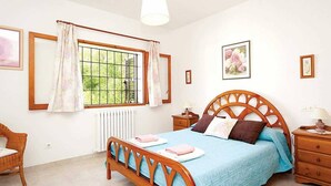 A bedroom with a wooden bed, a window with curtains, a bedside table, and a chair. - Casa para familias en Ibiza (Sant Josep de Sa Talaia)