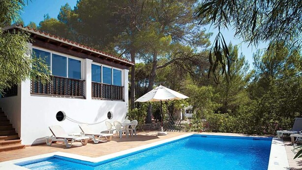A swimming pool with a white pool edge, a white building with a balcony, and a pergola with an umbrella. - Casa para familias en Ibiza (Sant Josep de Sa Talaia)