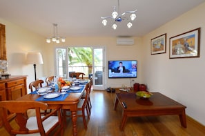 A dining area with a wooden table, chairs, a television, and a window with a view of palm trees. - Acogedor apartamento a 100 metros de la playa (Palmanova)