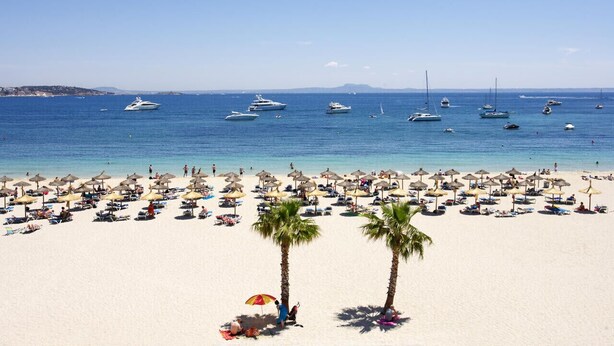 A beach with numerous umbrellas and sun loungers, a clear blue sea, and several boats in the background. - Acogedor apartamento a 100 metros de la playa (Palmanova)