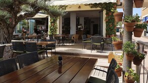 An outdoor cafe with wooden tables and chairs, surrounded by potted plants and a large tree. - Acogedor apartamento a 100 metros de la playa (Palmanova)