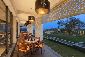 Un área de comedor al aire libre con mesas y sillas, vista al paisaje y un edificio al fondo.