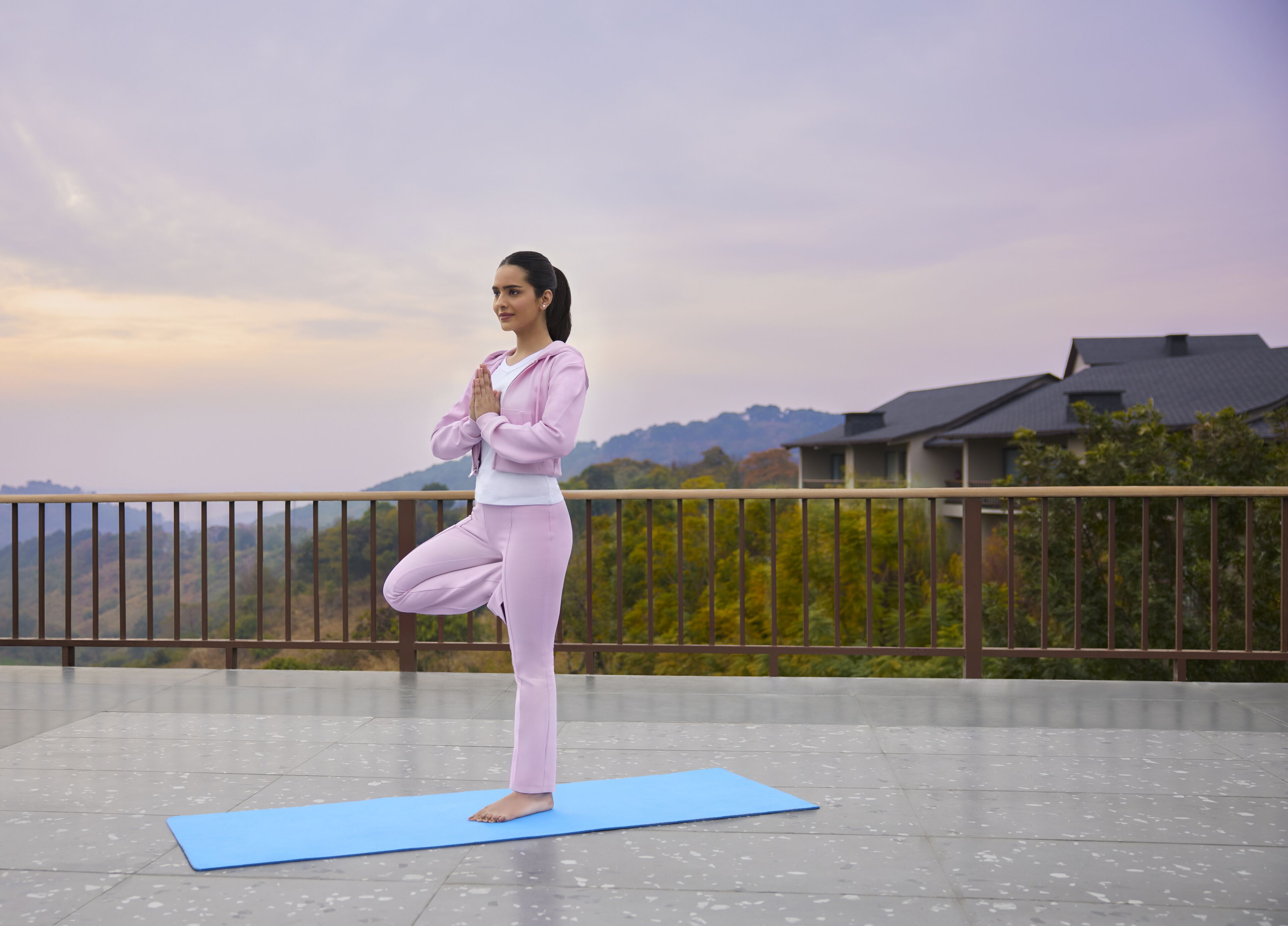 A person performing a yoga pose on a blue mat outdoors with a view of houses and trees in the background.
