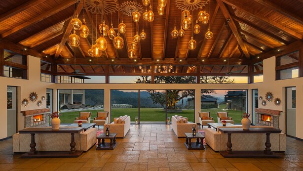 Amplio espacio interior con techo alto, varias lámparas colgantes y una chimenea. Varias zonas de estar con sofás y mesas, y ventanas grandes que ofrecen vistas a un jardín.