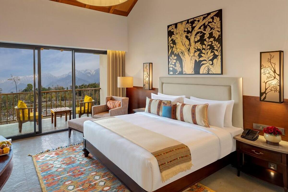 Quarto de hotel com uma cama grande, varanda com cadeiras, vista para montanhas e obras de arte nas paredes.