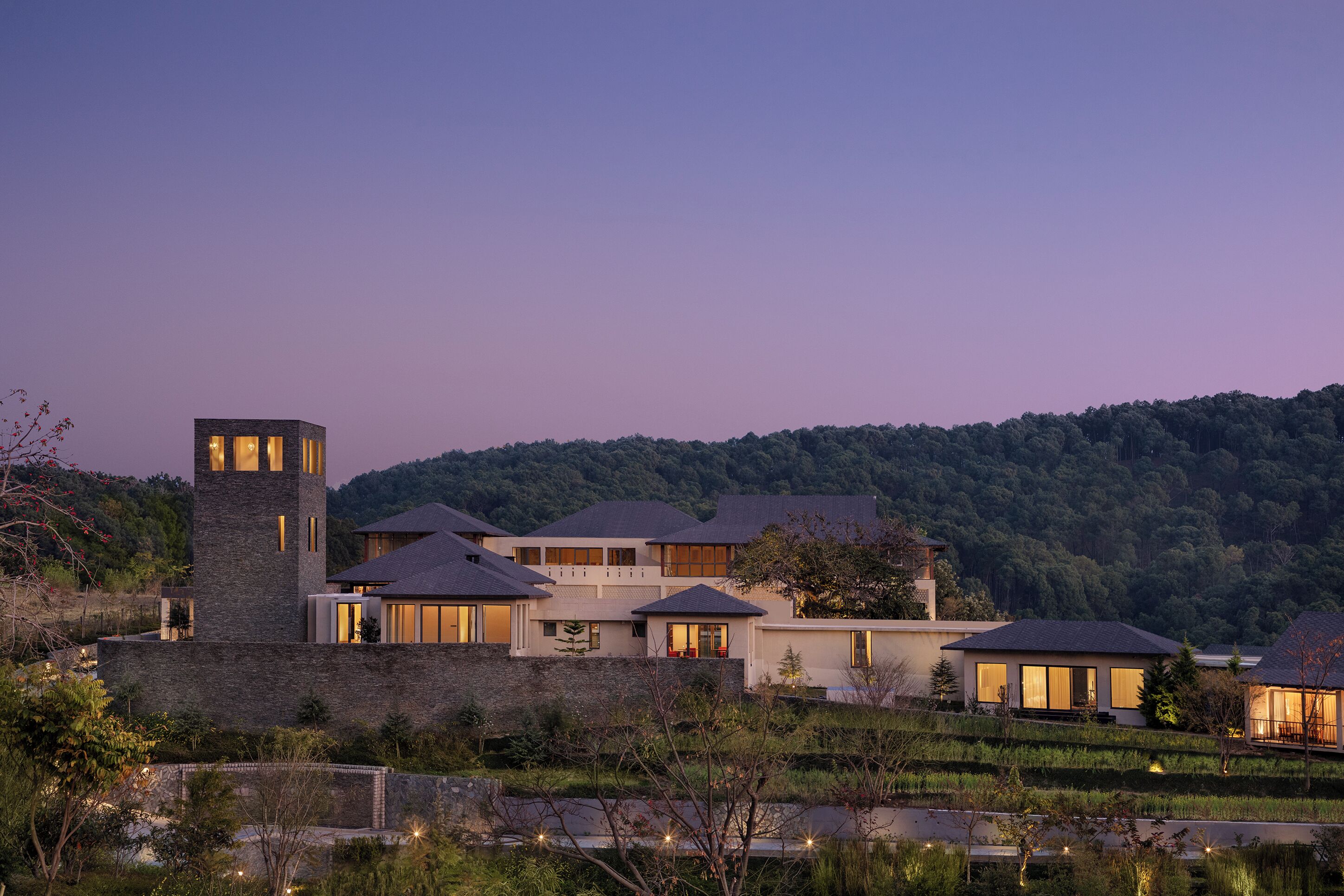 A modern multi-story house with a stone tower, surrounded by lush greenery and hills.