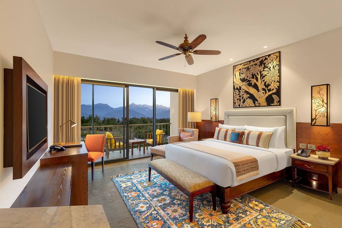 Quarto de hotel com uma cama grande, uma escrivaninha, uma cadeira, um ventilador de teto e vista para as montanhas.