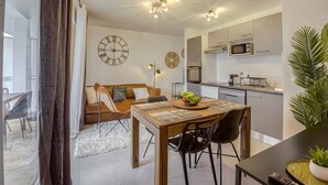 A modern kitchen with a microwave, oven, and sink. There is a dining area with a wooden table and chairs, and a sofa with cushions. A clock is on the wall, and a plant is on the counter.